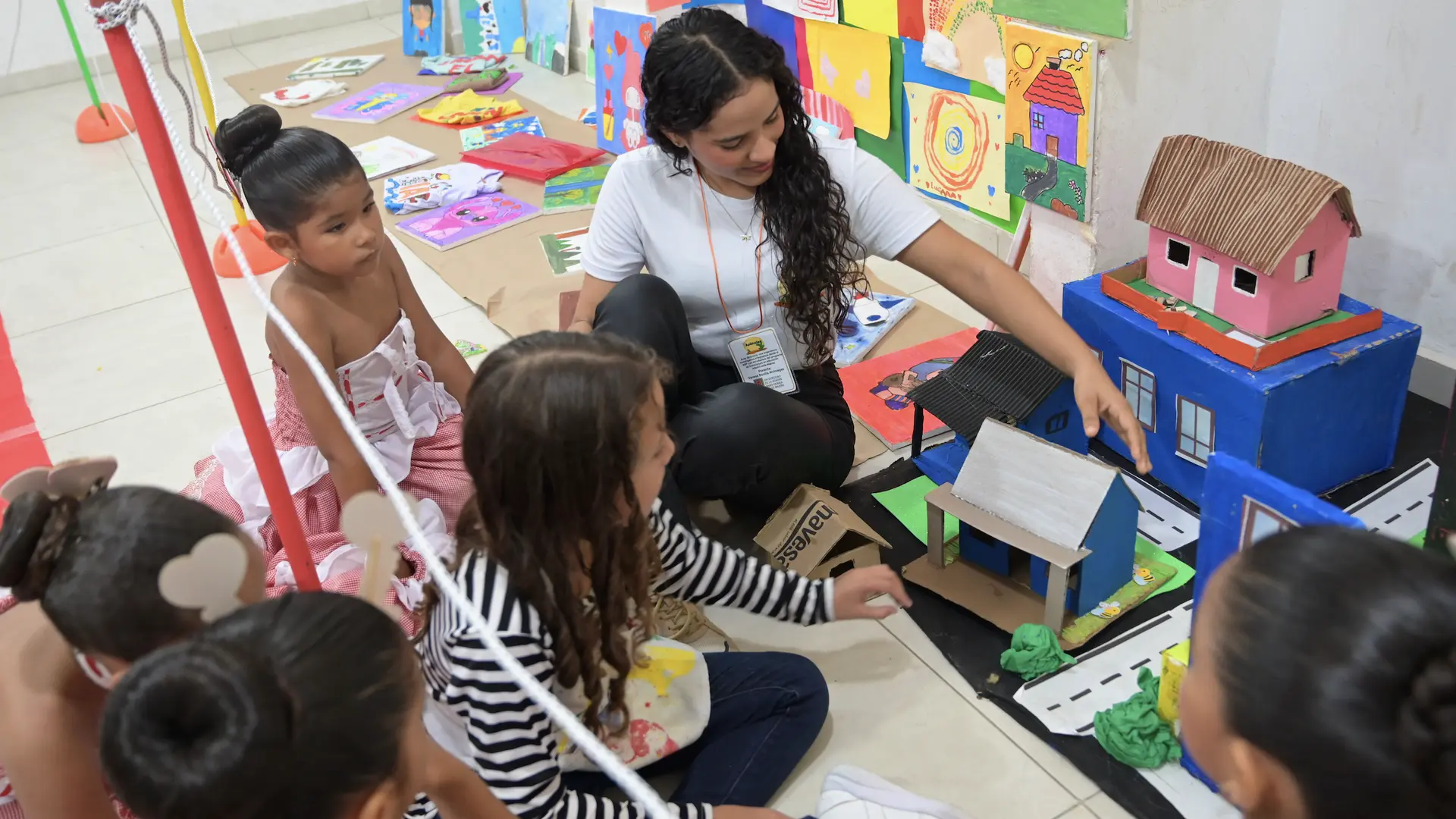 Una estudiante interactúa con un grupo de niñas pequeñas que observan una maqueta de casas de colores elaborada con cartón. La escena ocurre en un aula decorada con dibujos y pinturas infantiles colocadas en la pared y el suelo. Las niñas, algunas vestidas con trajes típicos, participan con atención en la actividad educativa.