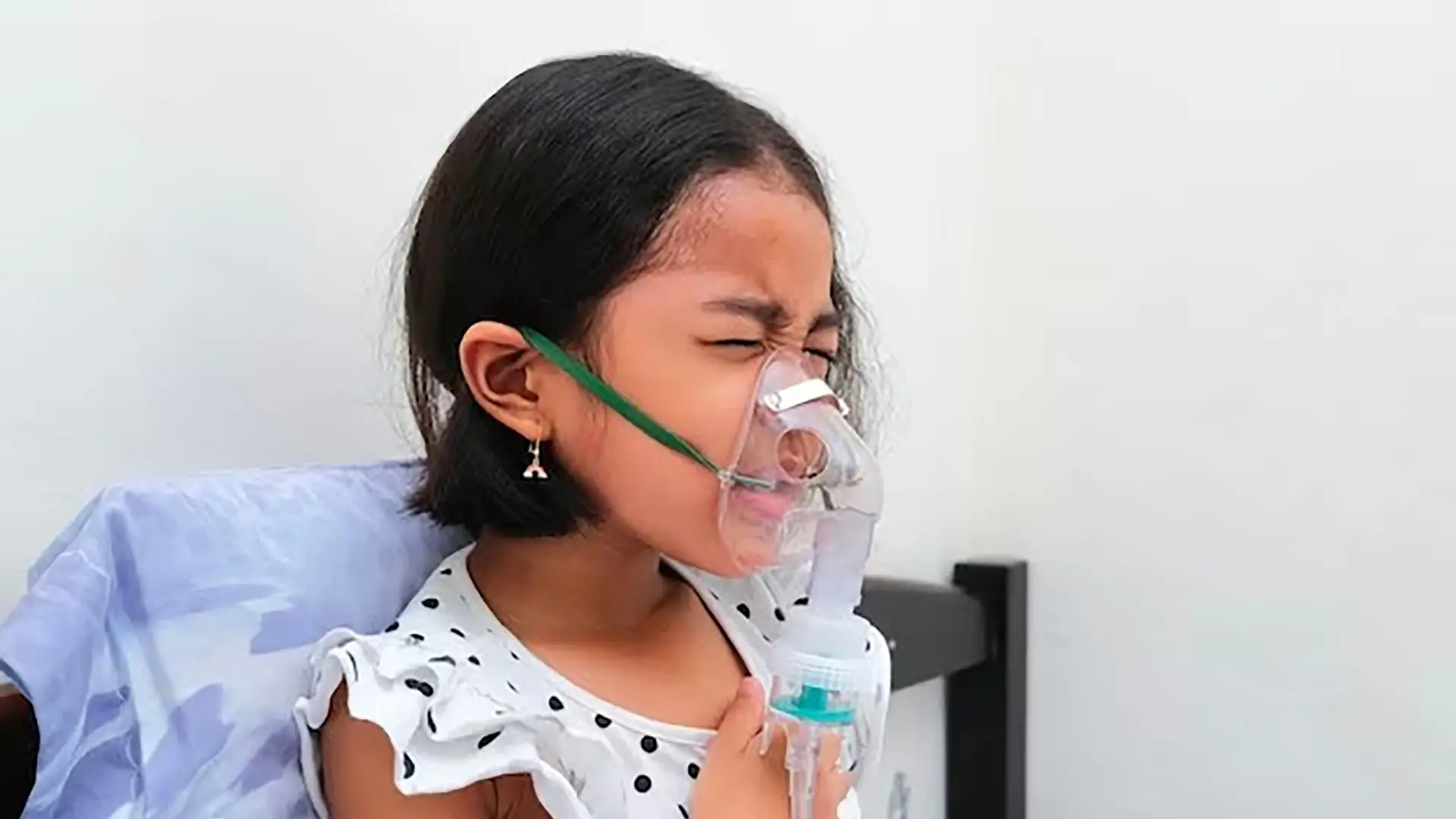 Niña pequeña sentada y usando una mascarilla de oxígeno conectada a un nebulizador. Tiene los ojos cerrados y una mano en el pecho, lo que sugiere que experimenta malestar o dificultad para respirar.