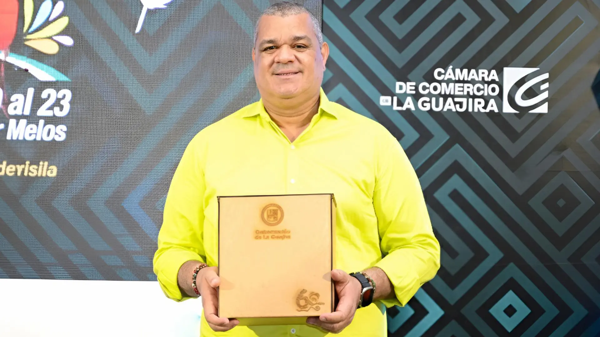Un hombre posa frente a un fondo con diseño geométrico y el logo de la Cámara de Comercio de La Guajira. Sostiene una caja con el sello de la Gobernación de La Guajira y luce camisa verde clara. El entorno corresponde a un evento institucional.