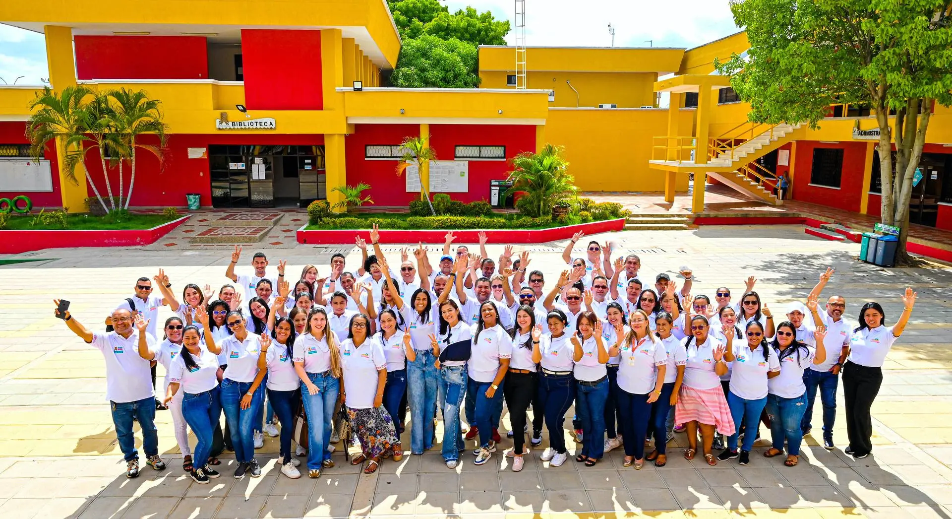 Grupo numeroso de personas adultas reunidas, posando y saludando con las manos levantadas. Todas visten camisetas blancas con logos institucionales. Al fondo se observan edificios de colores amarillo y rojo, áreas verdes y señalización de biblioteca, lo que indica un entorno universitario al aire libre, en este caso en Uniguajira Sede Maicao.