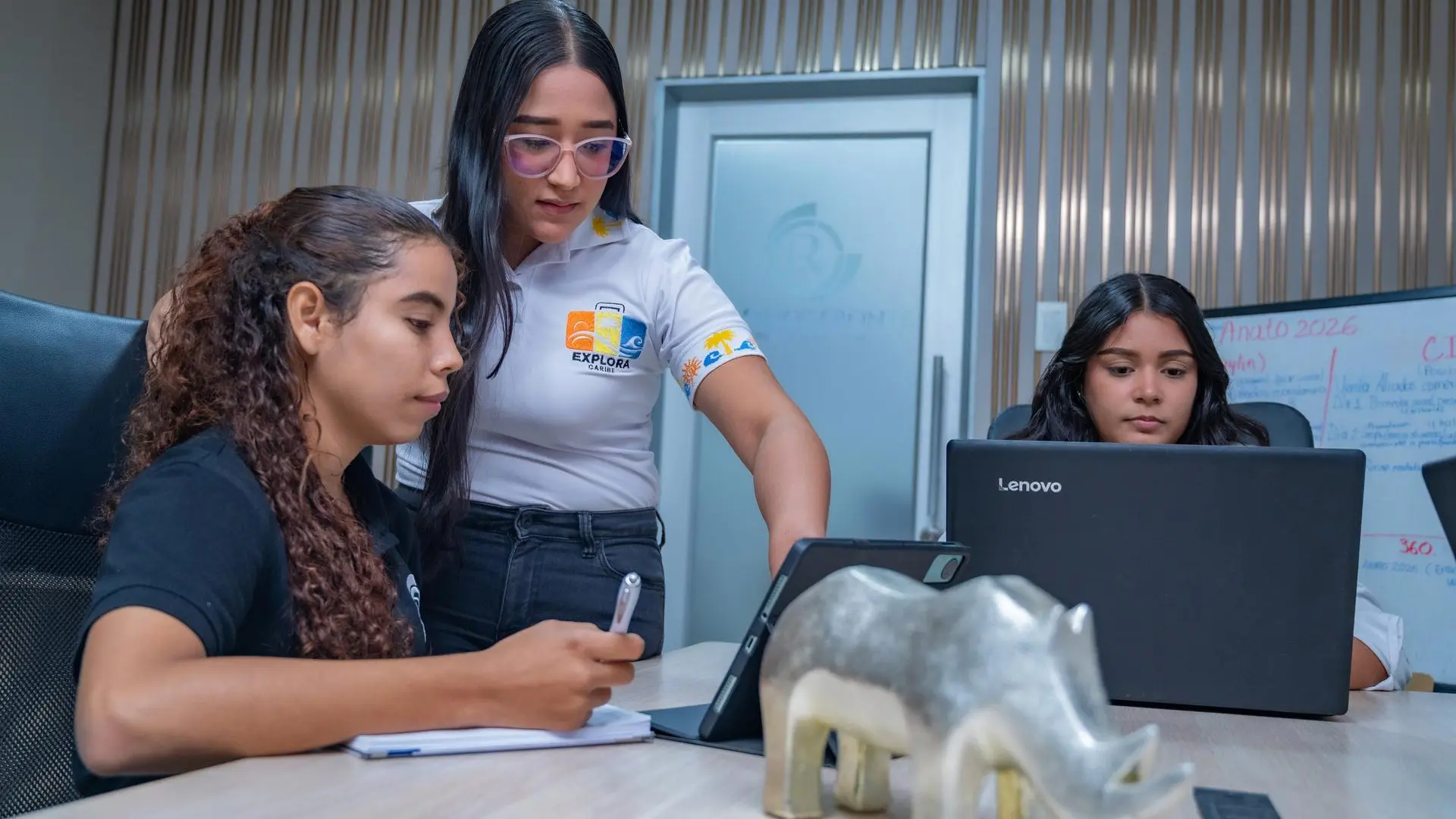 Tres mujeres trabajan en equipo alrededor de una mesa con portátil y tableta, revisando información en una oficina moderna.