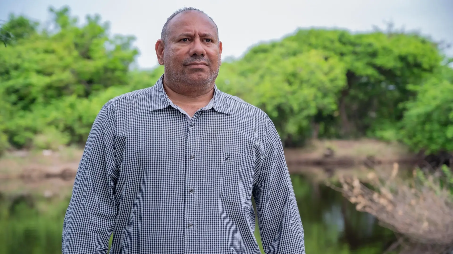 Retrato de medio cuerpo de un hombre posando con expresión serena en un paisaje natural con árboles verdes y un cuerpo de agua al fondo. Viste una camisa formal de manga larga color gris.