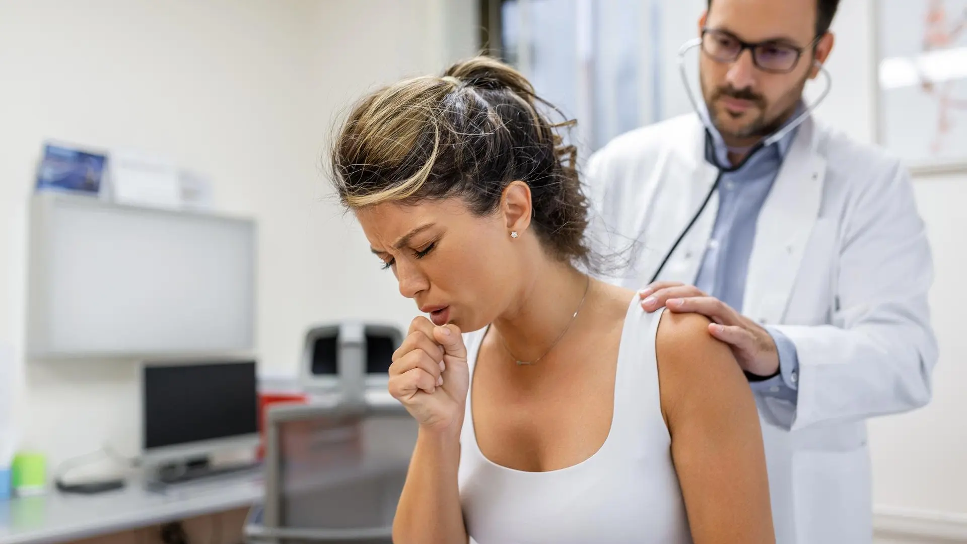 Mujer tosiendo durante una consulta médica mientras un doctor, con estetoscopio, ausculta sus pulmones en un consultorio.