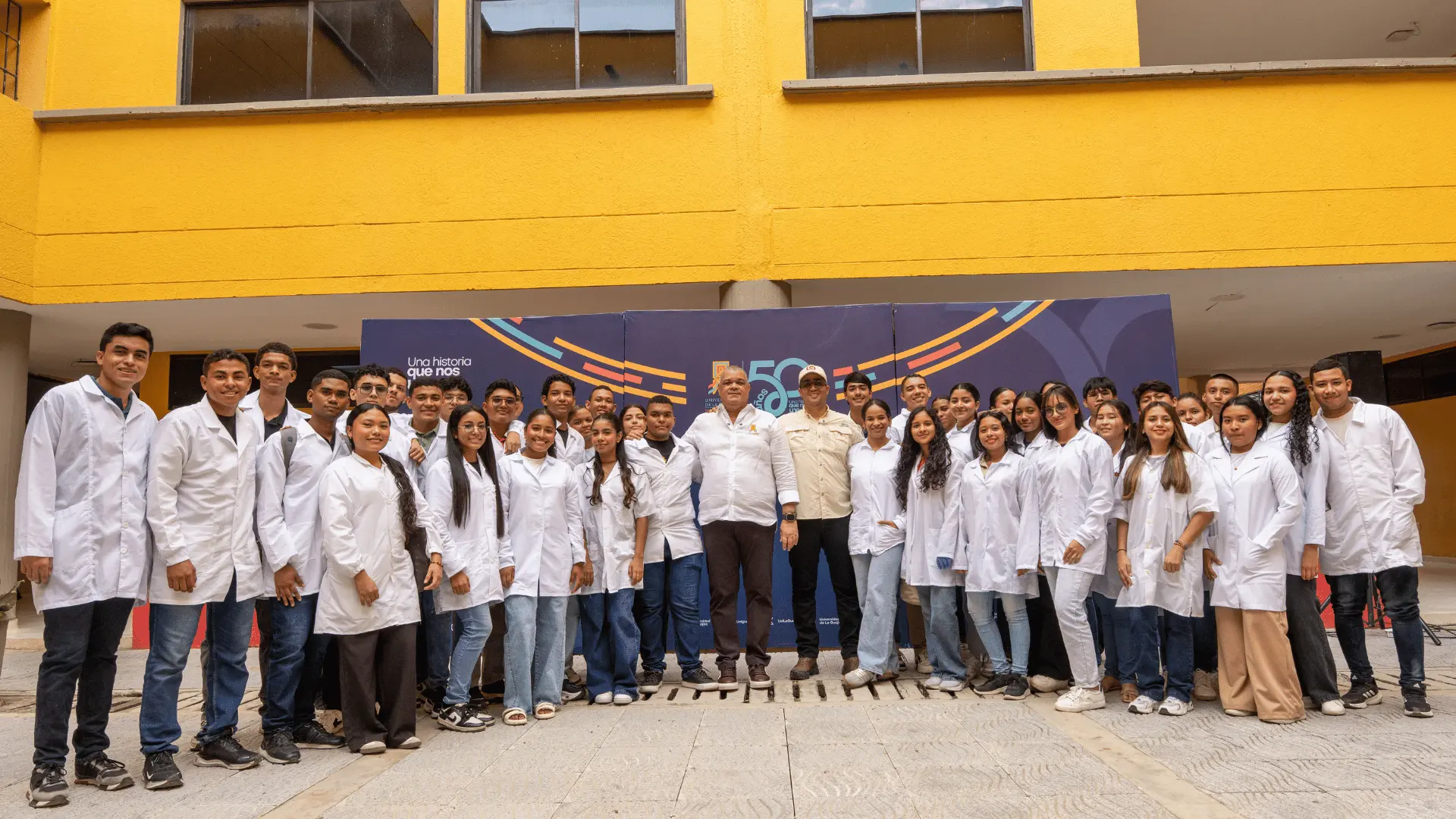 Grupo de estudiantes del programa de Medicina con batas blancas posa junto al rector y al gobernador de La Guajira frente a un panel institucional . La fotografía fue tomada en un espacio abierto con fachada amarilla de fondo, durante una actividad académica o visita oficial.
