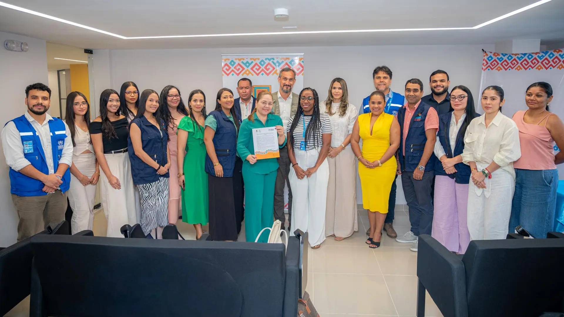 Un grupo de personas posa en fila dentro de una sala institucional durante un evento formal. En el centro, una mujer vestida de verde sostiene un documento firmado, rodeada por representantes y colaboradores de Acnur y Uniguajira. El ambiente refleja un acto de colaboración o firma de acuerdo, con banners institucionales al fondo y asientos en primer plano.