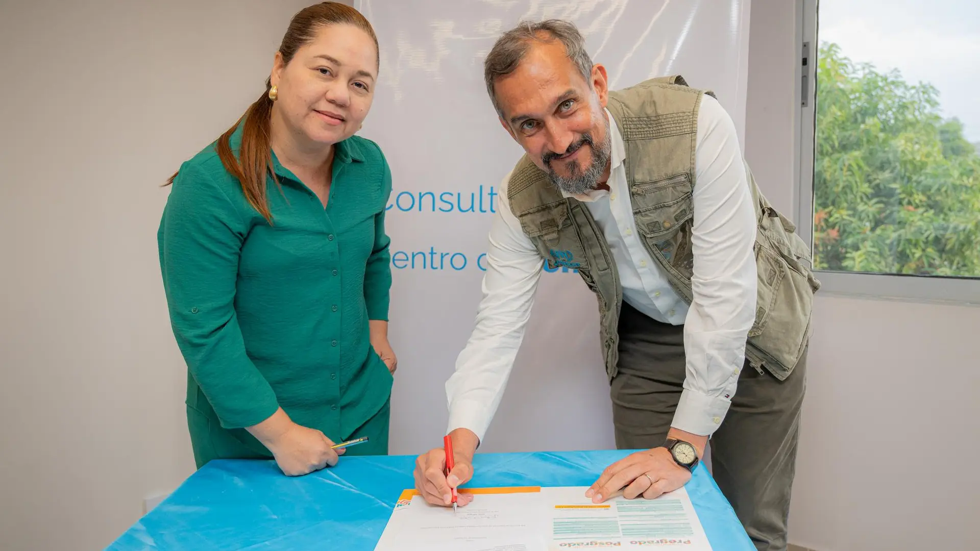 Dos personas están de pie mientras firman un documento, un hombre con chaleco beige se inclina para firmar con un bolígrafo rojo, mientras una mujer con blusa verde observa y sostiene un lápiz. El momento corresponde a la firma de un acuerdo en un espacio interior iluminado, con una ventana y un fondo institucional detrás.