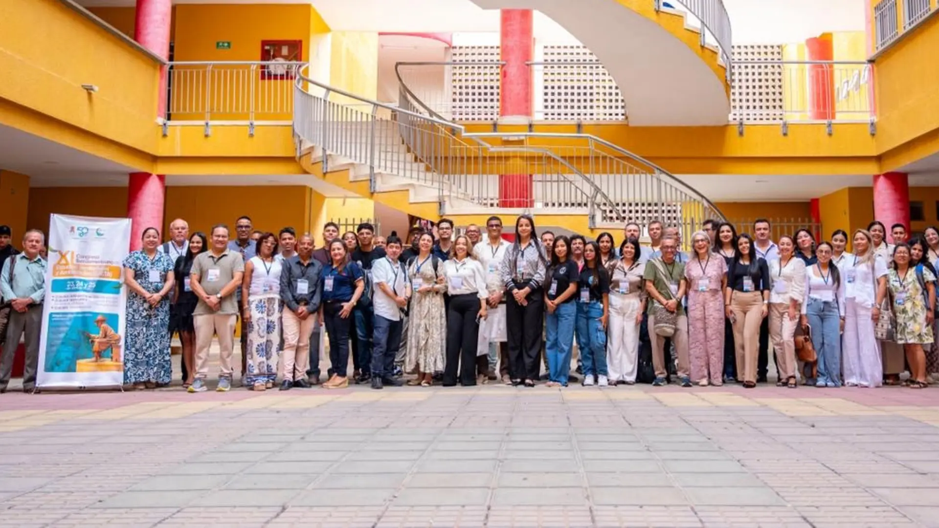 Grupo numeroso de personas posando en un patio institucional con escaleras y columnas de colores, durante un evento académico o encuentro profesional.