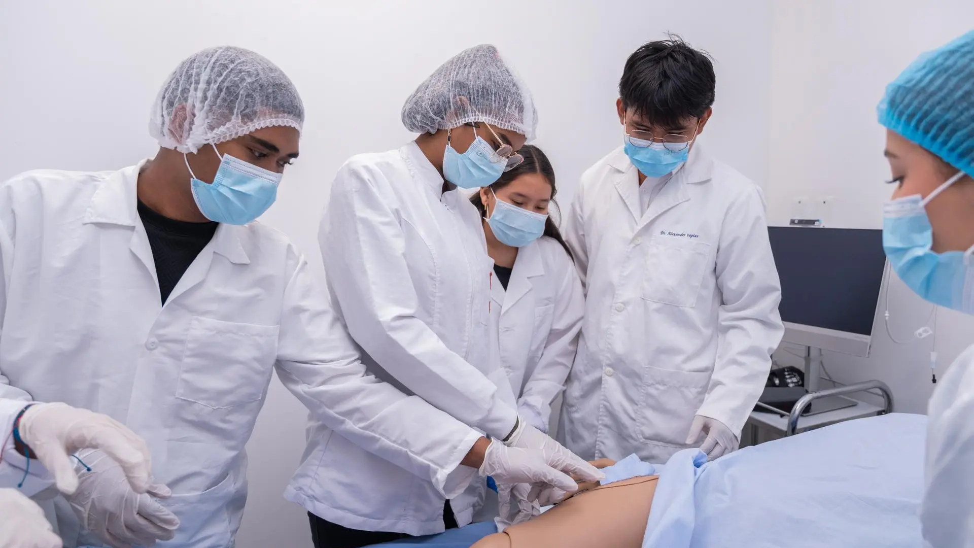 Grupo de estudiantes con batas blancas, gorros y tapabocas realiza una práctica médica en un laboratorio, trabajando sobre un maniquí de simulación mientras uno de ellos guía el procedimiento y los demás observan y colaboran.