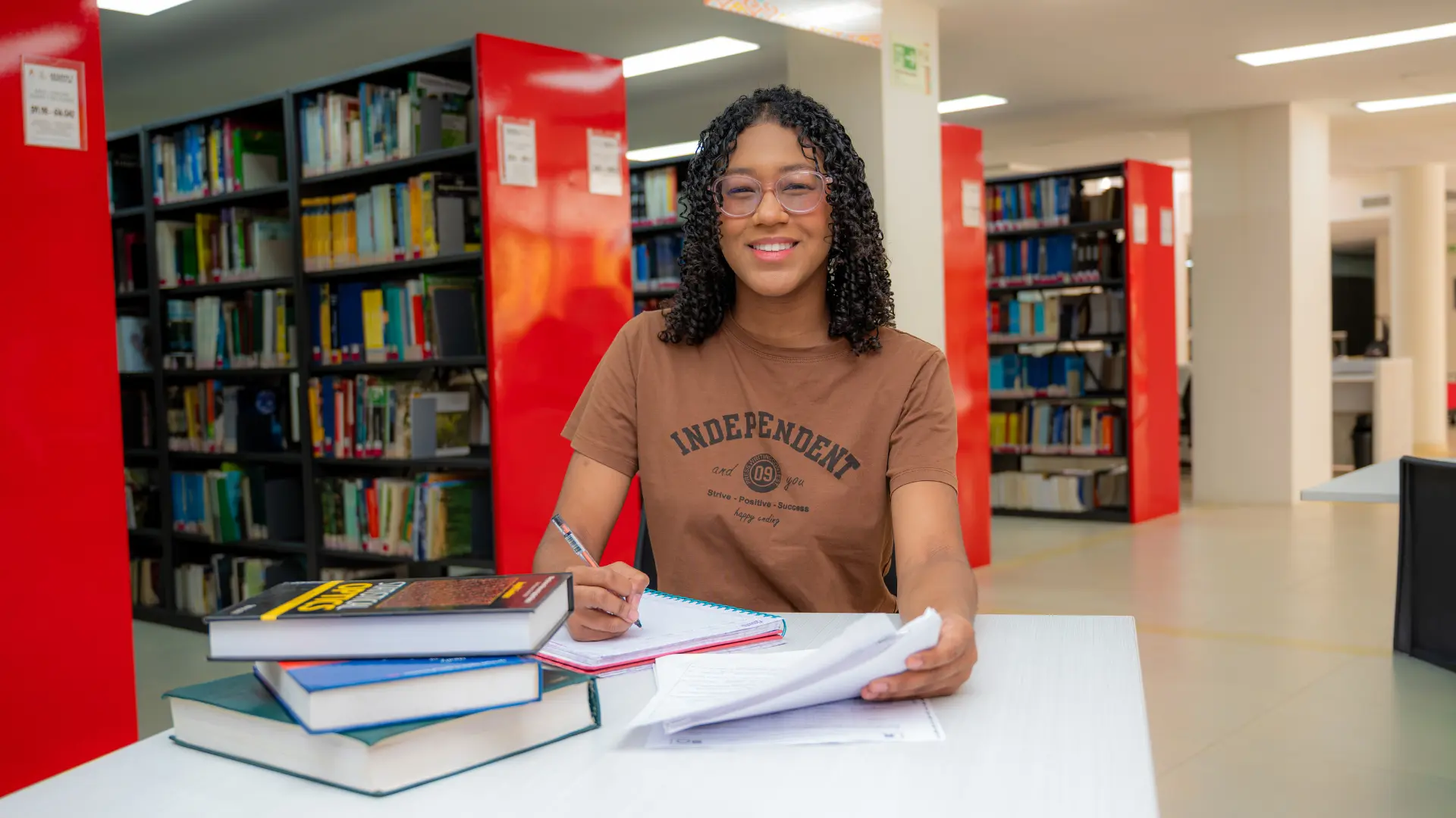 Estudiante sentada en una biblioteca, sonríe mientras escribe en un cuaderno, con varios libros apilados sobre la mesa y estanterías al fondo.