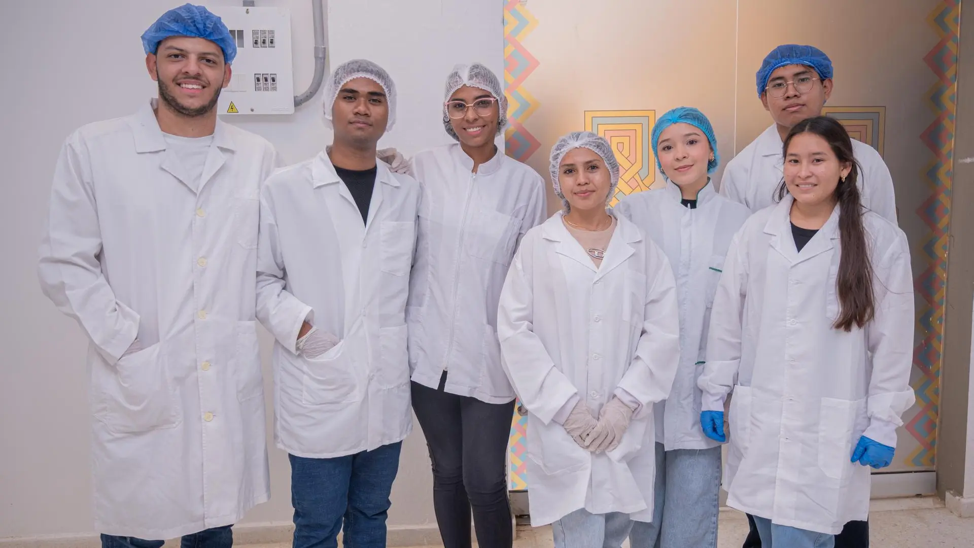 Grupo de siete estudiantes en un entorno tipo laboratorio o área de prácticas, vestidos con batas blancas, gorros de protección y algunos con guantes. Están de pie, uno junto al otro, mirando a la cámara y sonriendo. El espacio incluye paredes claras, un panel eléctrico y una puerta con detalles de colores al fondo, lo que sugiere un ambiente académico o de formación técnica.