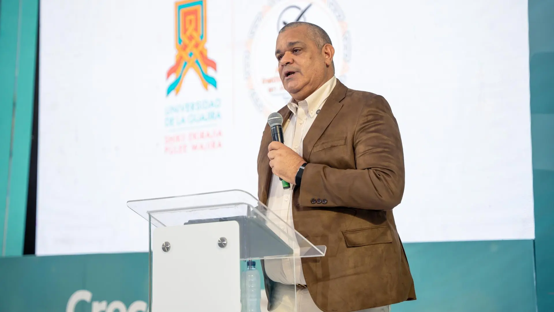Hombre adulto ofrece una intervención pública desde un atril transparente, sosteniendo un micrófono. Viste chaqueta marrón y camisa clara. Al fondo se observa una pantalla con el logo institucional de la Universidad de La Guajira, en un escenario de evento académico o institucional.