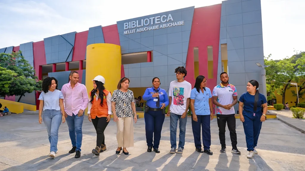 Nueve personas caminan juntas y conversan en un espacio interior amplio y moderno con columnas rojas, representando diversidad de profesiones y edades.