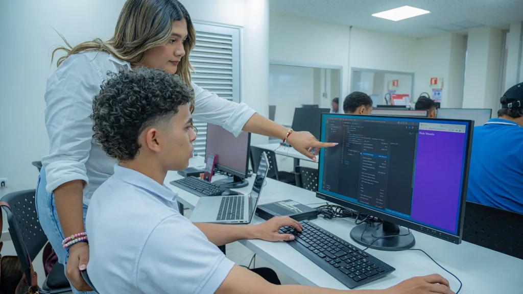 Dos jóvenes estudiantes en un laboratorio de informática analizan líneas de código en un monitor de pantalla plana, mientras la joven señala un punto específico de la programación. El entorno es un aula moderna con iluminación brillante, equipada con teclados, computadoras portátiles y otros estudiantes trabajando al fondo.