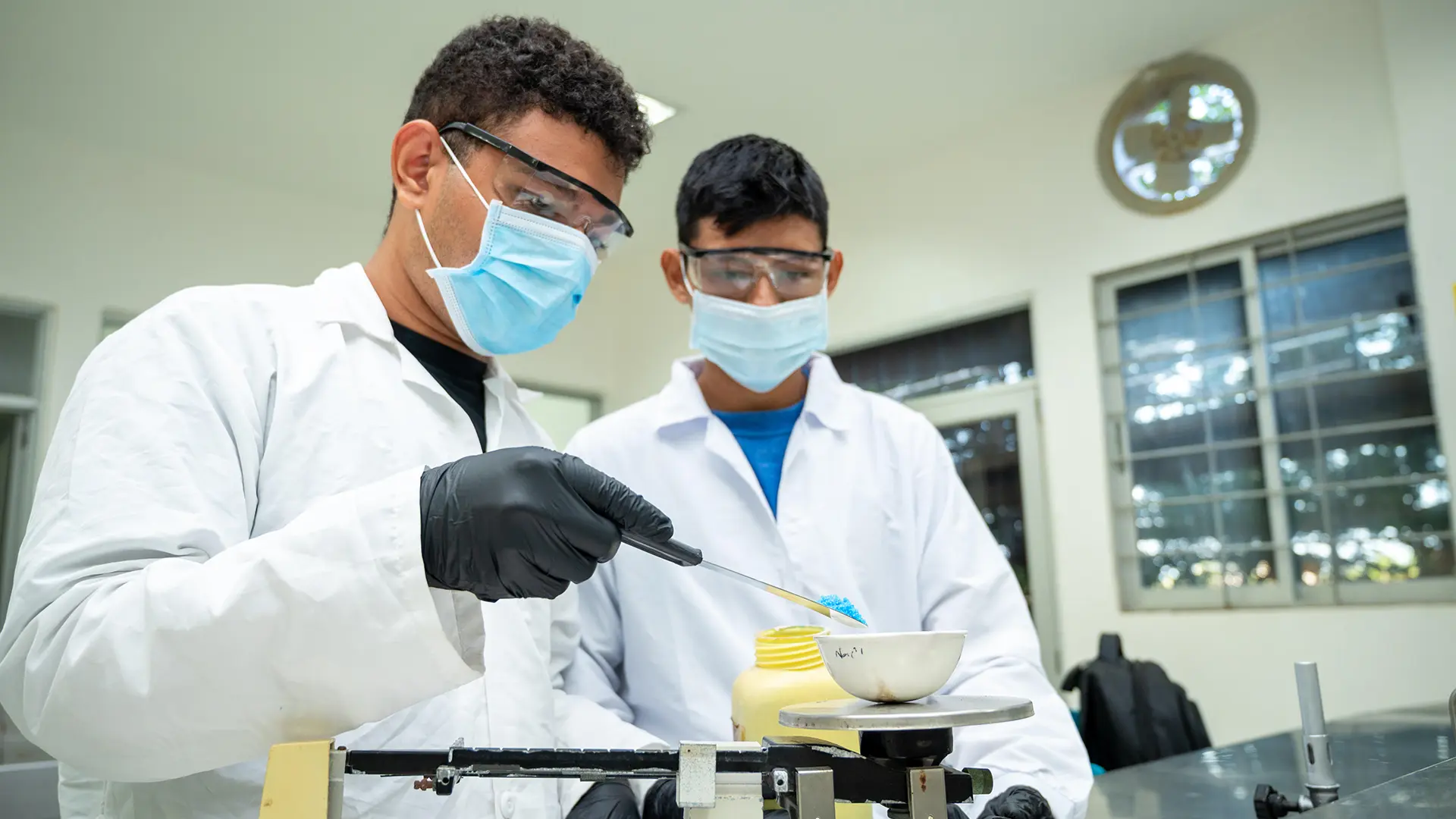 Dos estudiantes realizan una práctica de laboratorio utilizando implementos de bioseguridad, mientras manipulan una muestra con instrumentos de medición en un entorno científico universitario.