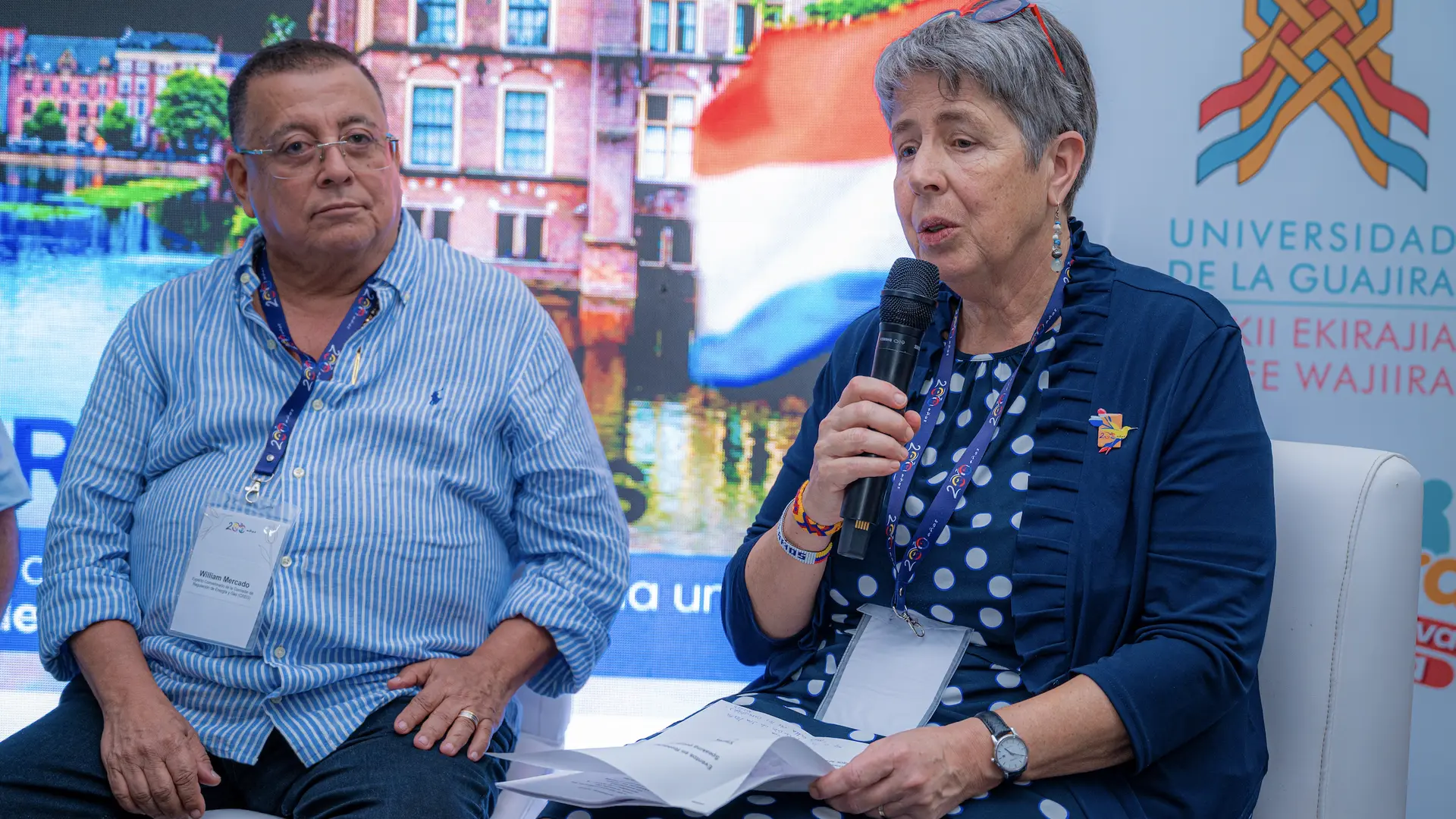 Una mujer con gafas sobre el cabello y vestido azul de puntos blancos habla por un micrófono durante un panel de discusión. A su lado, un hombre con camisa de rayas azules escucha atentamente mientras ambos están sentados frente a un fondo informativo. A la derecha se observa el logotipo de la Universidad de La Guajira.