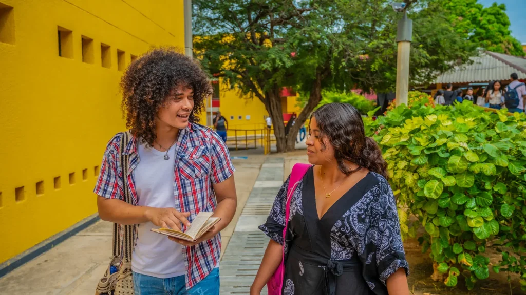 Dos estudiantes conversan mientras caminan por un campus universitario rodeado de árboles y edificios amarillos.