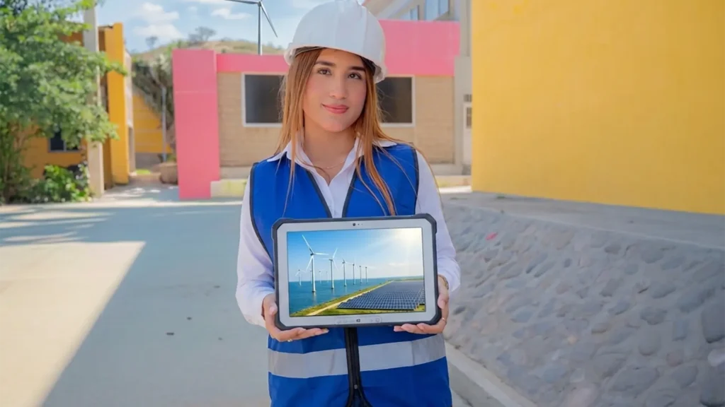 Una mujer joven con casco de seguridad blanco y chaleco reflectante azul sostiene una tableta que muestra un parque de energía eólica y solar. Se encuentra en un entorno interior de la Universidad de La Guajira con colores institucionales y una pequeña turbina eólica al fondo.