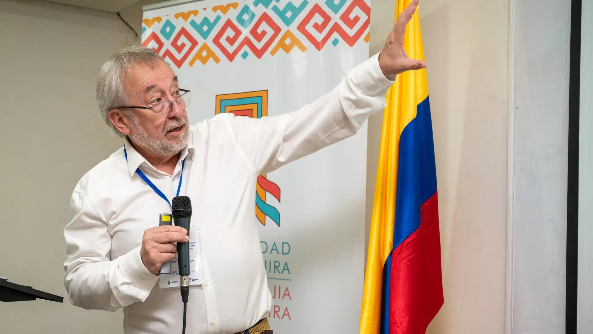 Conferencista participa en una presentación académica dentro de un auditorio. El expositor, vestido con camisa blanca y portando una escarapela institucional, sostiene un micrófono mientras señala una proyección fuera de cámara. Detrás de él se observa un pendón institucional con motivos gráficos inspirados en la cultura wayuu y, a un costado, la bandera de Colombia. La imagen corresponde a una intervención durante un evento académico o científico.