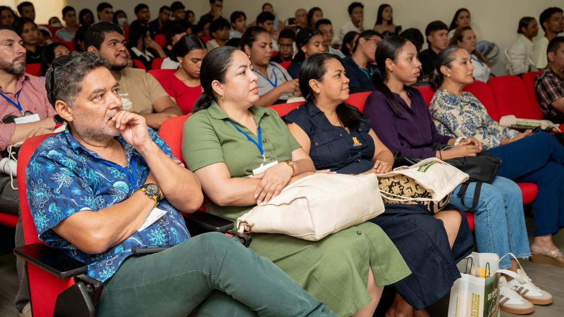 Auditorio con decenas de asistentes sentados en sillas rojas durante una conferencia o evento académico. En primer plano, cinco personas observan atentamente la presentación fuera de cámara; algunas portan escarapelas institucionales y llevan bolsos o materiales de apoyo. Al fondo se aprecia una audiencia diversa conformada por estudiantes, docentes y profesionales, concentrados en la actividad. El espacio cuenta con iluminación interior y paredes claras, propias de una sala de conferencias universitaria.
