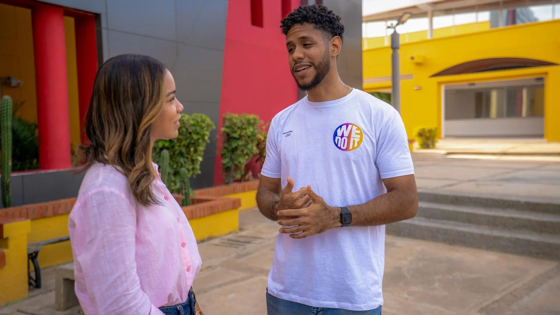 Un hombre con camiseta blanca que lleva el logotipo "WE DO IT" habla y gesticula con las manos frente a una mujer con camisa rosa en un patio exterior, con edificios de colores llamativos al fondo.