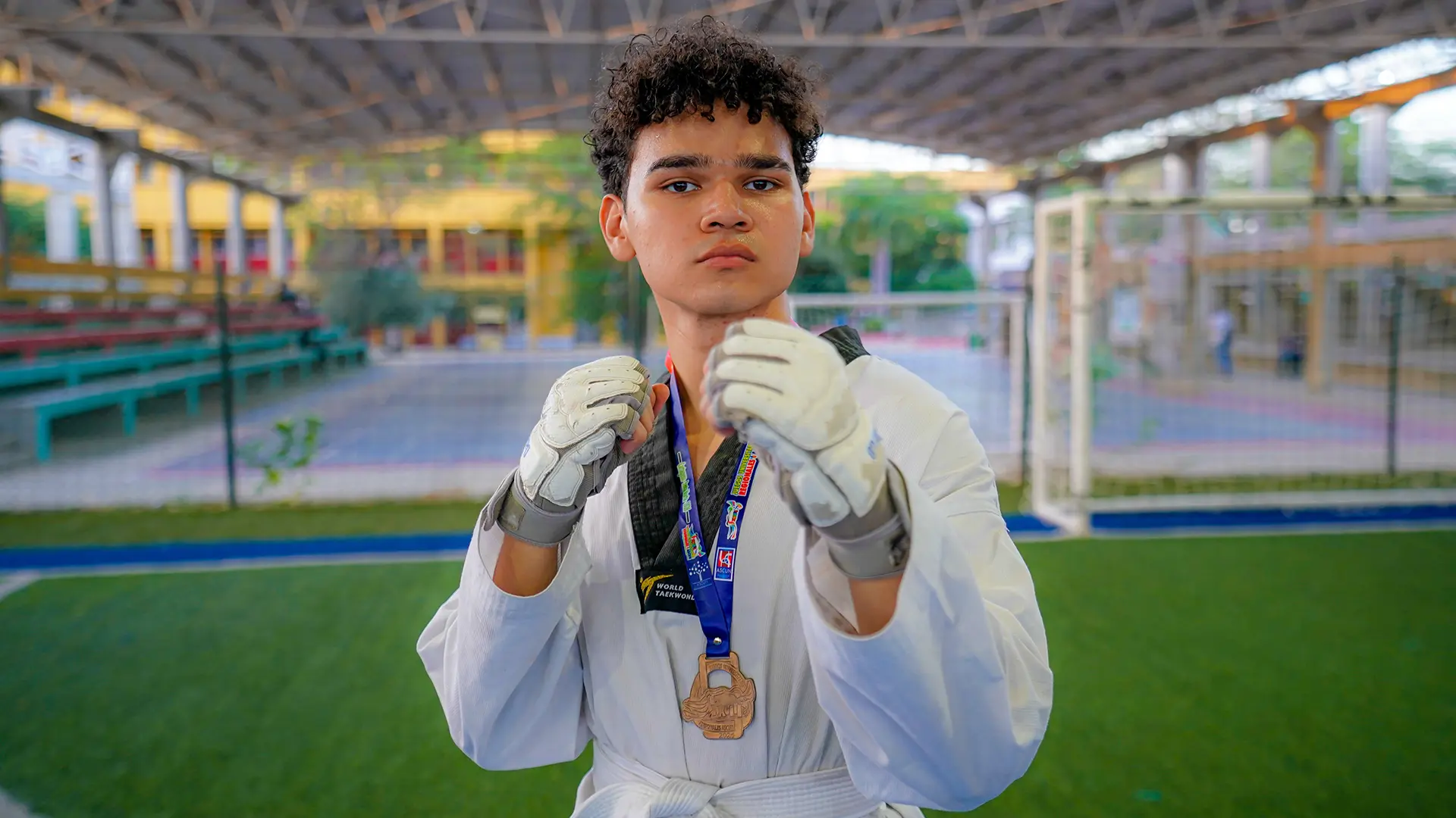 Fotografía de un estudiante en una cancha deportiva cubierta de la Universidad de La Guajira. El joven viste uniforme blanco de taekwondo, cinturón blanco y guantes de protección, mientras adopta una postura de combate con los puños levantados frente a la cámara. Lleva una medalla colgada al cuello, símbolo de un logro deportivo. Al fondo se observan las graderías, una portería y las instalaciones del campus desenfocadas. La imagen transmite disciplina, esfuerzo y dedicación al deporte universitario.