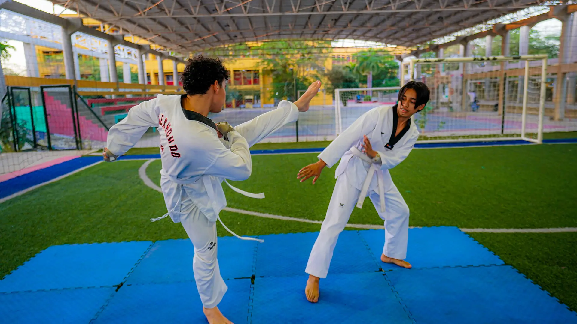 Fotografía de dos estudiantes practicando taekwondo en una cancha deportiva cubierta de la Universidad de La Guajira. Ambos visten uniforme blanco de taekwondo con cinturón blanco y se encuentran sobre colchonetas azules. Uno de los estudiantes ejecuta una patada lateral mientras el otro adopta una postura defensiva para esquivarla. Al fondo se observan una portería, graderías y las instalaciones del campus. La imagen refleja trabajo en equipo, disciplina, concentración y formación deportiva dentro del entorno universitario.