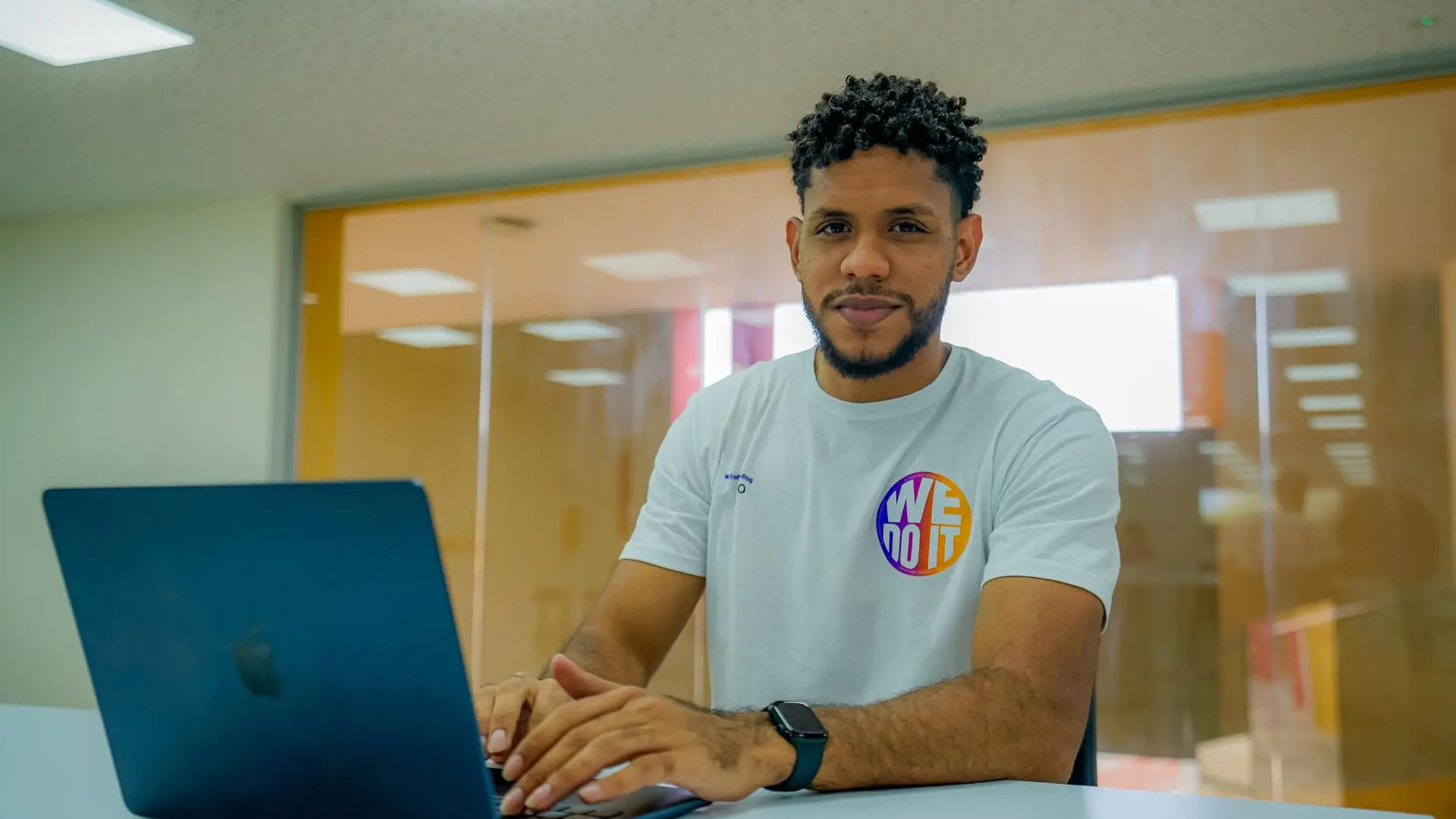 Un hombre con cabello rizado y barba mira fijamente a la cámara, con las manos sobre el teclado de una computadora portátil abierta en el primer plano a la izquierda. Lleva una camiseta blanca con el lema "WE DO IT" y un reloj inteligente en su muñeca izquierda, sentado frente a una pared de vidrio en una oficina moderna.