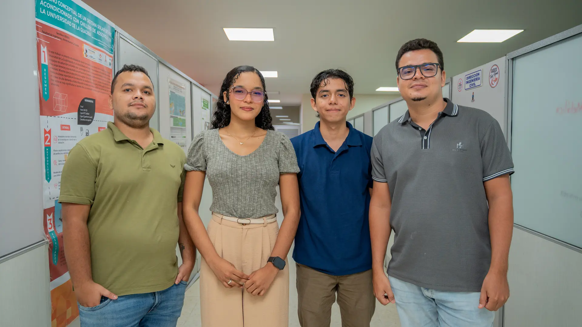 Cuatro jóvenes, tres hombres y una mujer, posan de pie en el pasillo de una institución educativa junto a un póster de diseño conceptual de la Universidad de La Guajira.