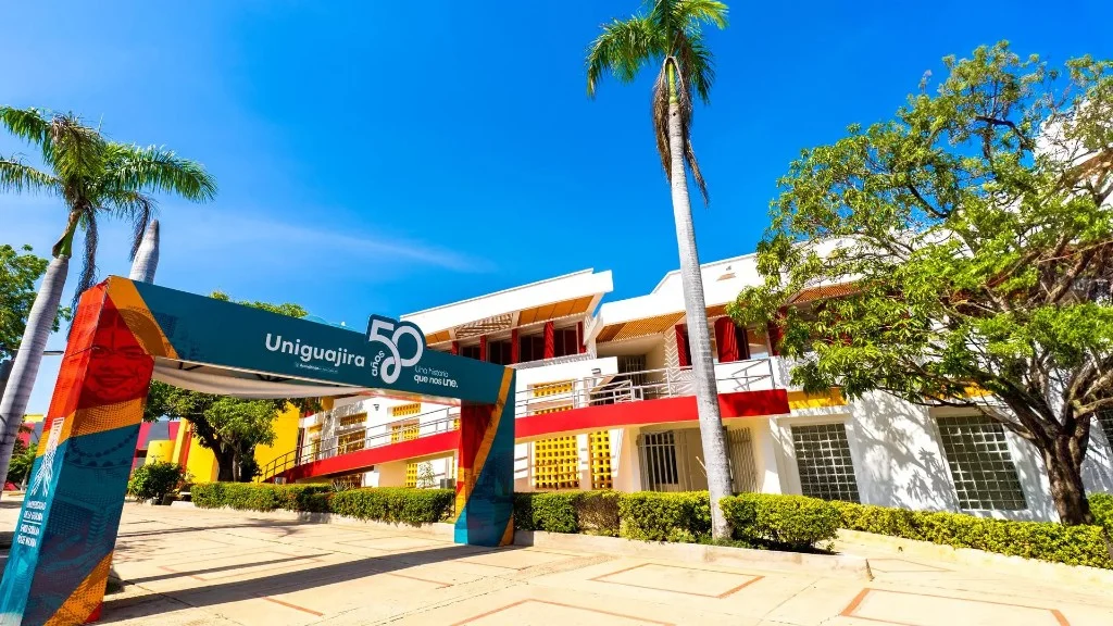 Vista exterior del campus de la Universidad de La Guajira, con el arco institucional conmemorativo de los 50 años en primer plano, edificios académicos al fondo, palmeras y zonas verdes bajo un cielo despejado.