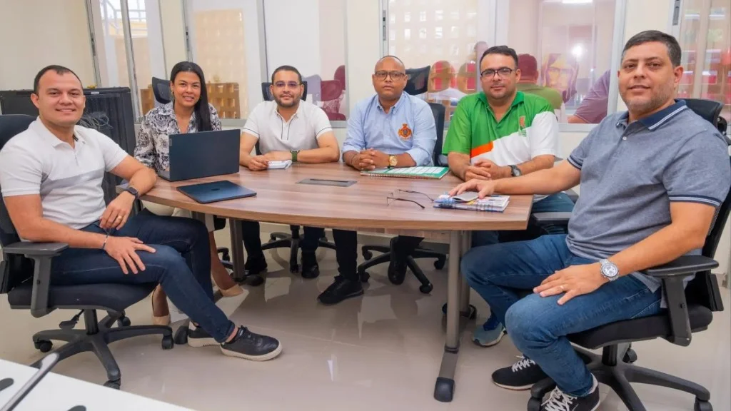 Seis integrantes del equipo de trabajo de la Universidad de La Guajira participan en una reunión alrededor de una mesa de conferencias, con computadores y documentos de trabajo, en un ambiente colaborativo dentro de una oficina institucional.