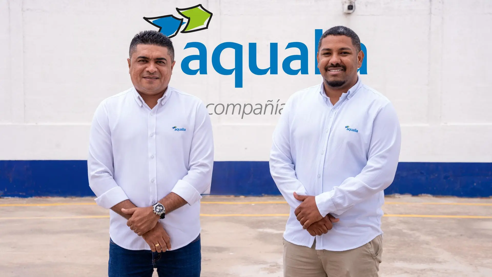 Dos colaboradores de Aqualia posan de pie frente al logotipo de la empresa en las instalaciones corporativas. Ambos visten camisas blancas con el logotipo institucional y mantienen una postura profesional mientras miran hacia la cámara.