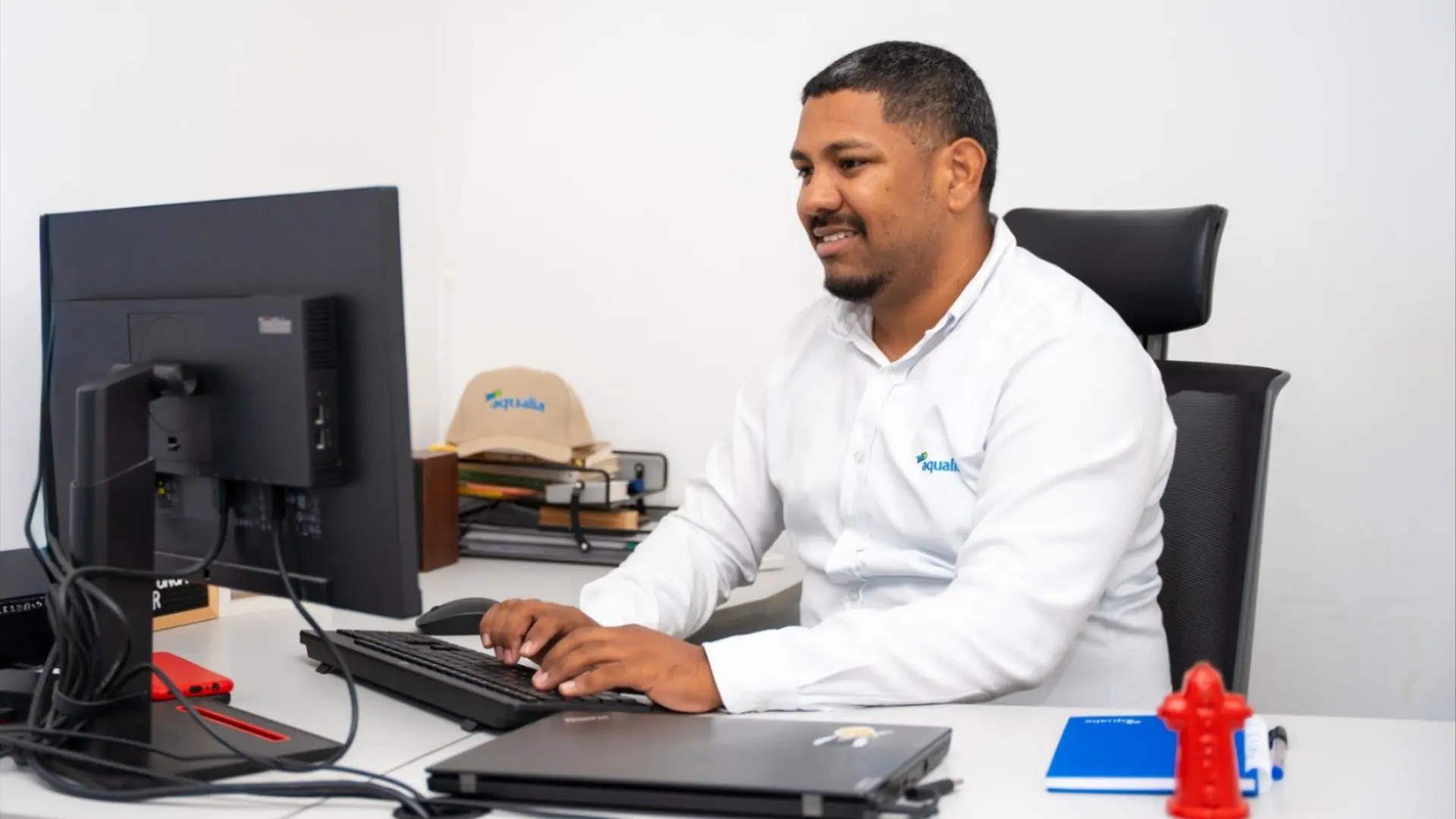 Colaborador de Aqualia sentado en su puesto de trabajo, utilizando un computador de escritorio en una oficina moderna. Viste una camisa blanca con el logotipo de la empresa y trabaja frente a un monitor, con un portátil, teclado y elementos de oficina sobre el escritorio.