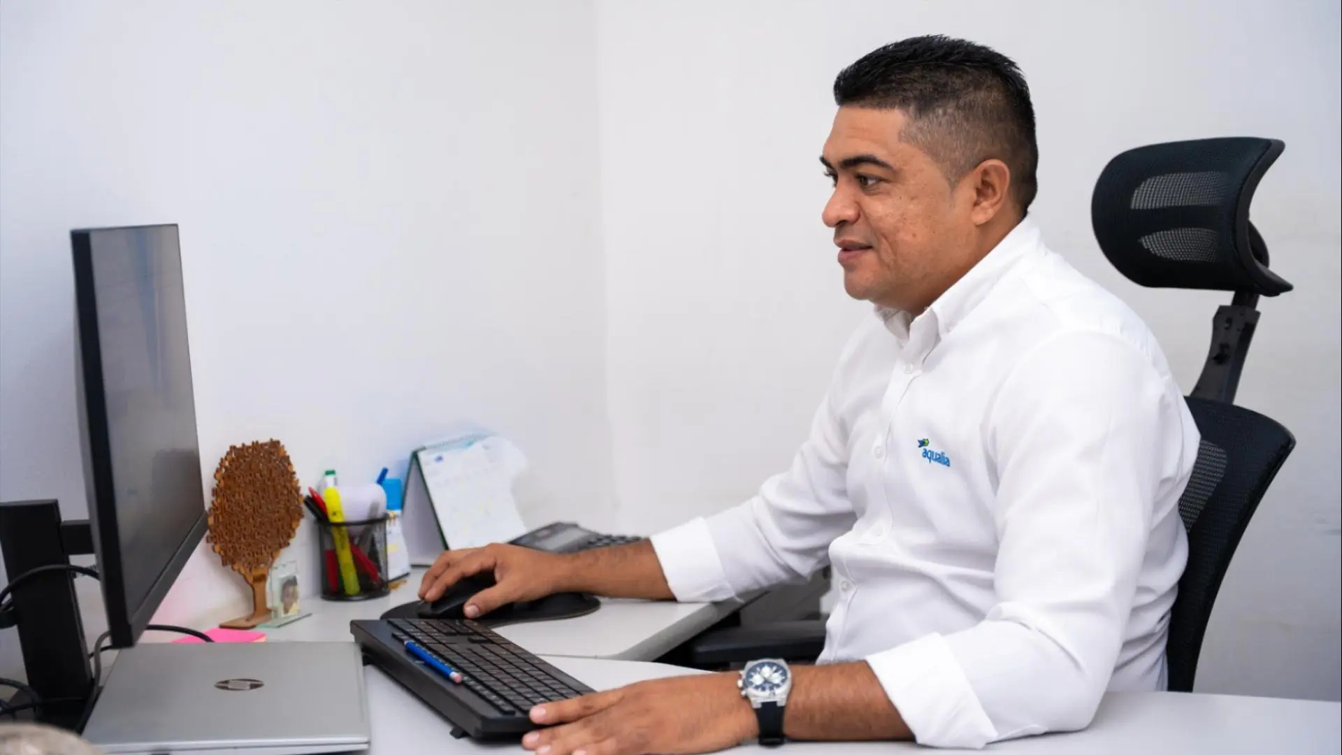 Colaborador de Aqualia trabajando frente a un computador de escritorio en una oficina. Lleva una camisa blanca con el logotipo de la empresa y sonríe mientras utiliza el teclado. En el escritorio se observan un portátil, una libreta y otros implementos de trabajo
