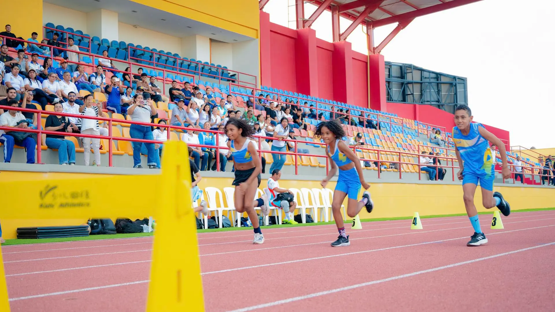 Niños y niñas participan en una carrera de atletismo en una pista de tartán rojo, vistiendo uniformes deportivos azules. En primer plano se observan conos amarillos que delimitan los carriles. Al fondo, las graderías del estadio están ocupadas por espectadores.