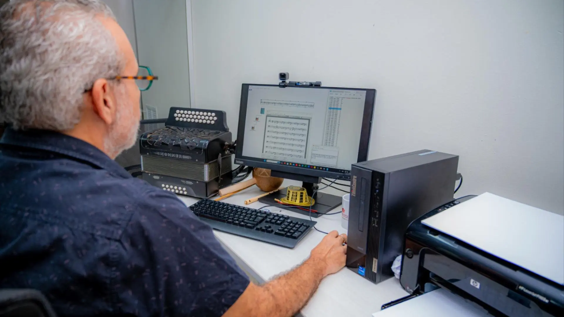 Hombre sentado frente a un computador, trabajando con un software de análisis musical. A su lado se encuentra un acordeón y, sobre el escritorio, otros instrumentos y elementos relacionados con la música