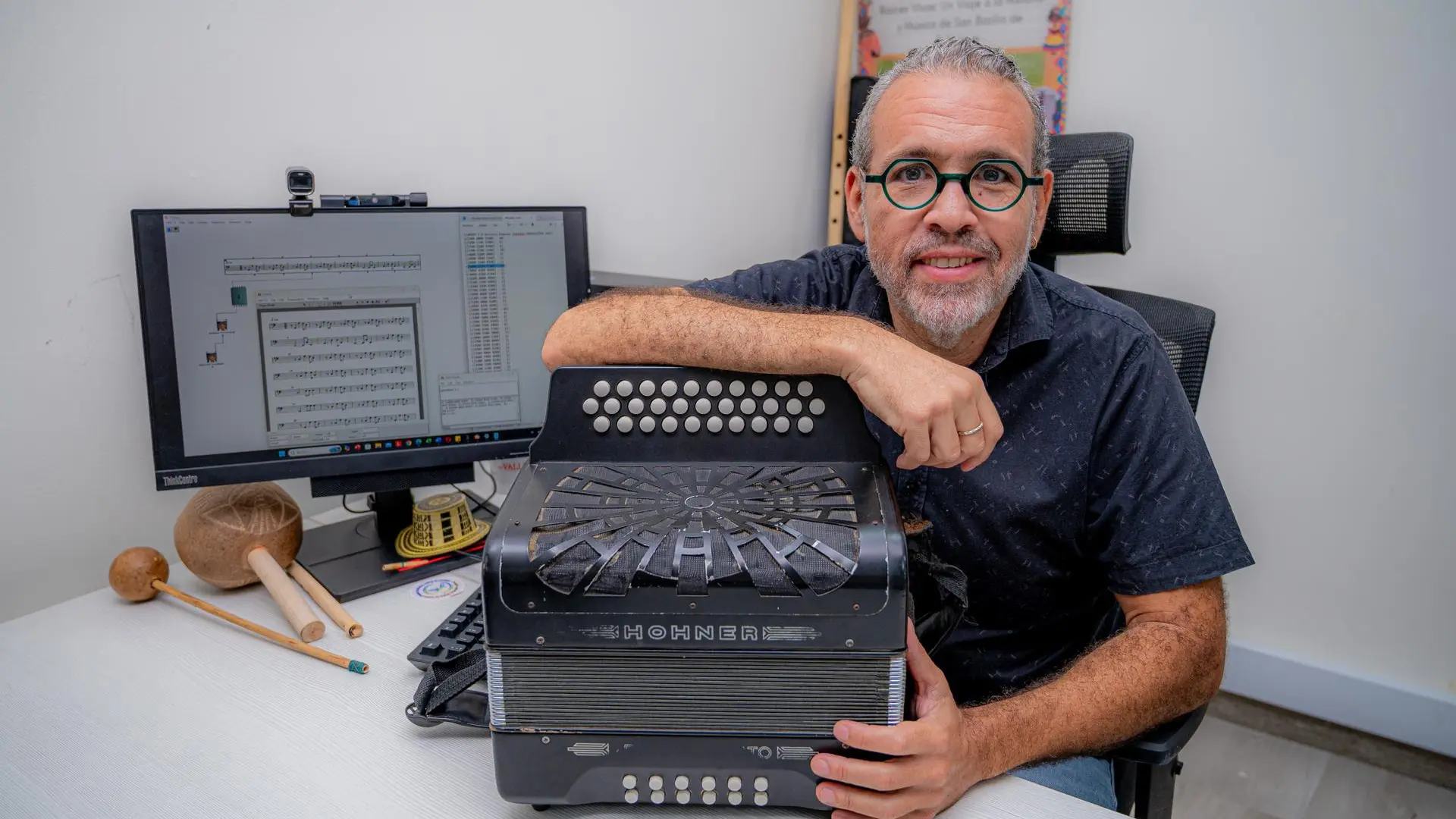 Hombre sentado frente a un computador y sosteniendo un acordeón, en un espacio de trabajo donde también se observan instrumentos musicales tradicionales.