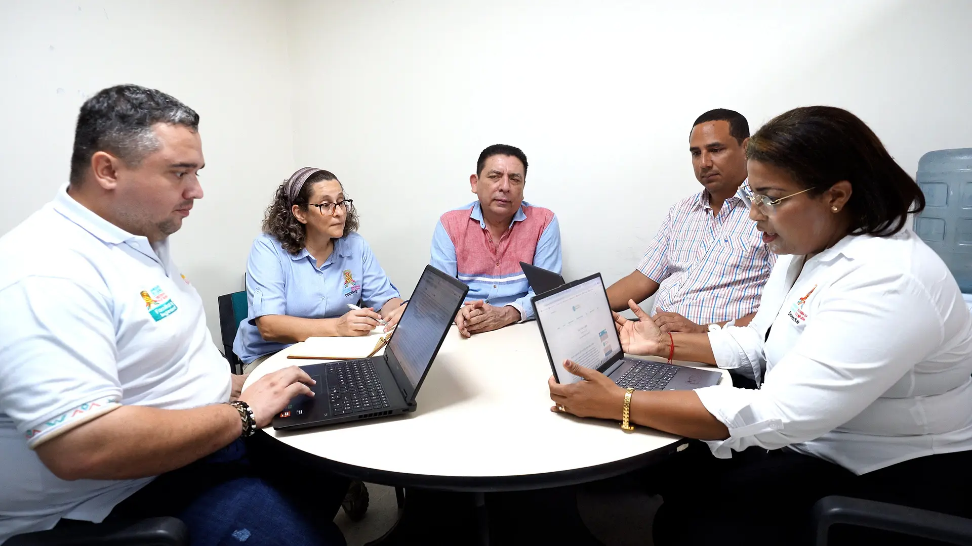 Cinco personas participan en una reunión de trabajo alrededor de una mesa redonda en una oficina. Tres utilizan computadores portátiles mientras una mujer toma apuntes en una libreta y otra persona expone información desde su portátil. Los asistentes mantienen una conversación en un ambiente colaborativo, con una pared blanca de fondo y un dispensador de agua al costado derecho.