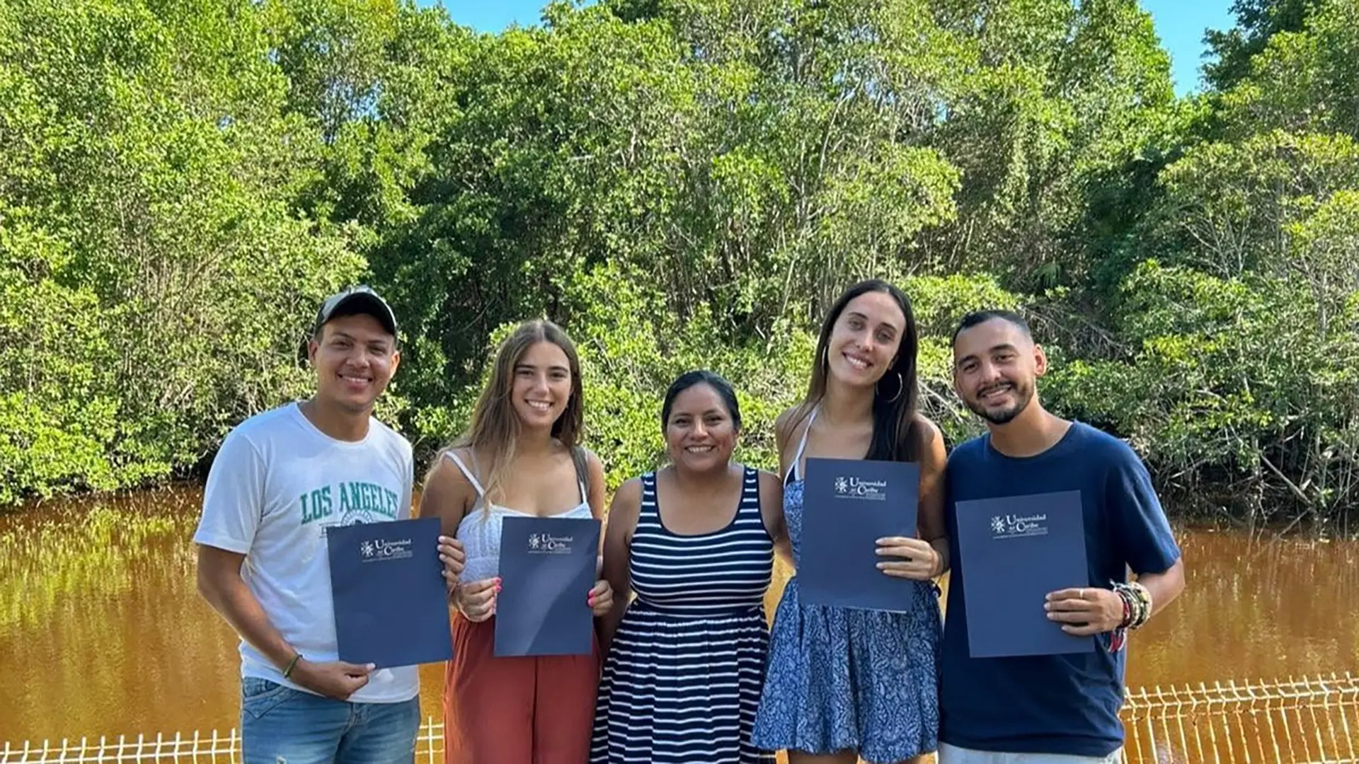 Grupo de estudiantes y una mujer posan al aire libre frente a un entorno natural, mientras sostienen documentos de movilidad académica.