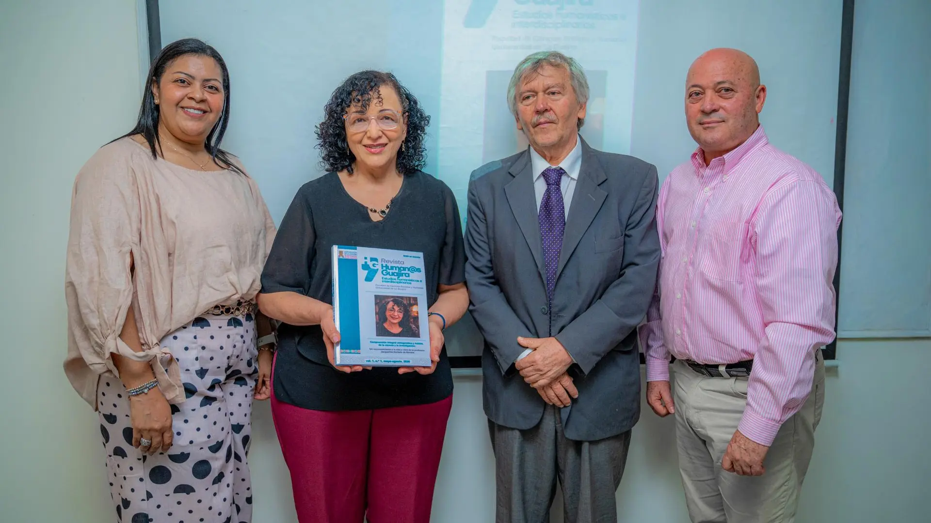 Cuatro personas posan durante la presentación de la revista Human@s Guajira. En el centro, una mujer sostiene un ejemplar impreso de la publicación, mientras las demás personas permanecen a su lado frente a una pantalla donde se proyecta la portada de la revista, en un acto académico o institucional.