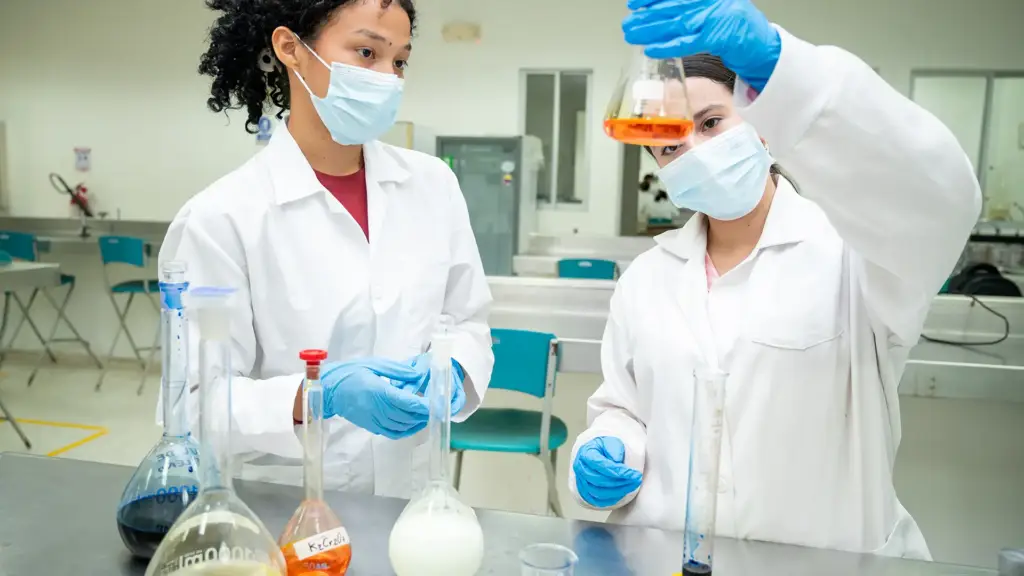 En un laboratorio de química, dos estudiantes con batas blancas y mascarillas quirúrgicas azules están frente a un banco de trabajo de acero inoxidable con cristalería variada. Una de ellas sostiene un matraz aforado con un líquido naranja y la otra la observa.