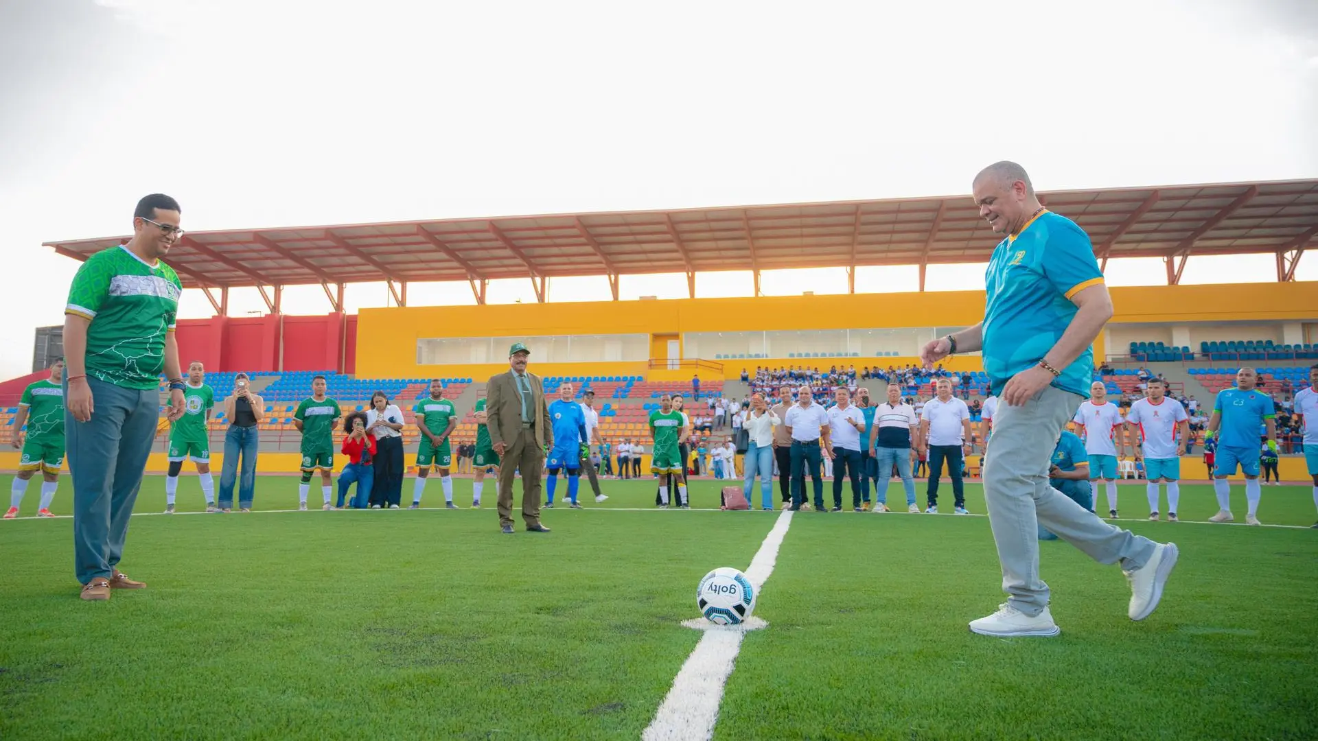 Personas reunidas en una cancha de fútbol durante la inauguración del escenario deportivo. En primer plano, el rector de Uniguajira y el gobernador del departamento se disponen a realizar el saque inicial frente a un balón ubicado sobre la línea central, mientras jugadores, autoridades y asistentes observan la actividad.