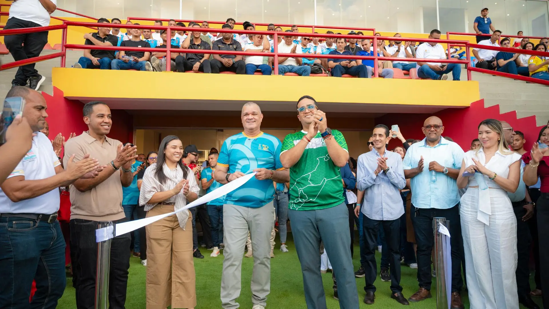 Personas reunidas en el escenario deportivo durante el acto protocolario de inauguración. En el centro, varios representantes sostienen y cortan una cinta blanca, mientras asistentes ubicados en las graderías y alrededor del lugar aplauden y acompañan la ceremonia