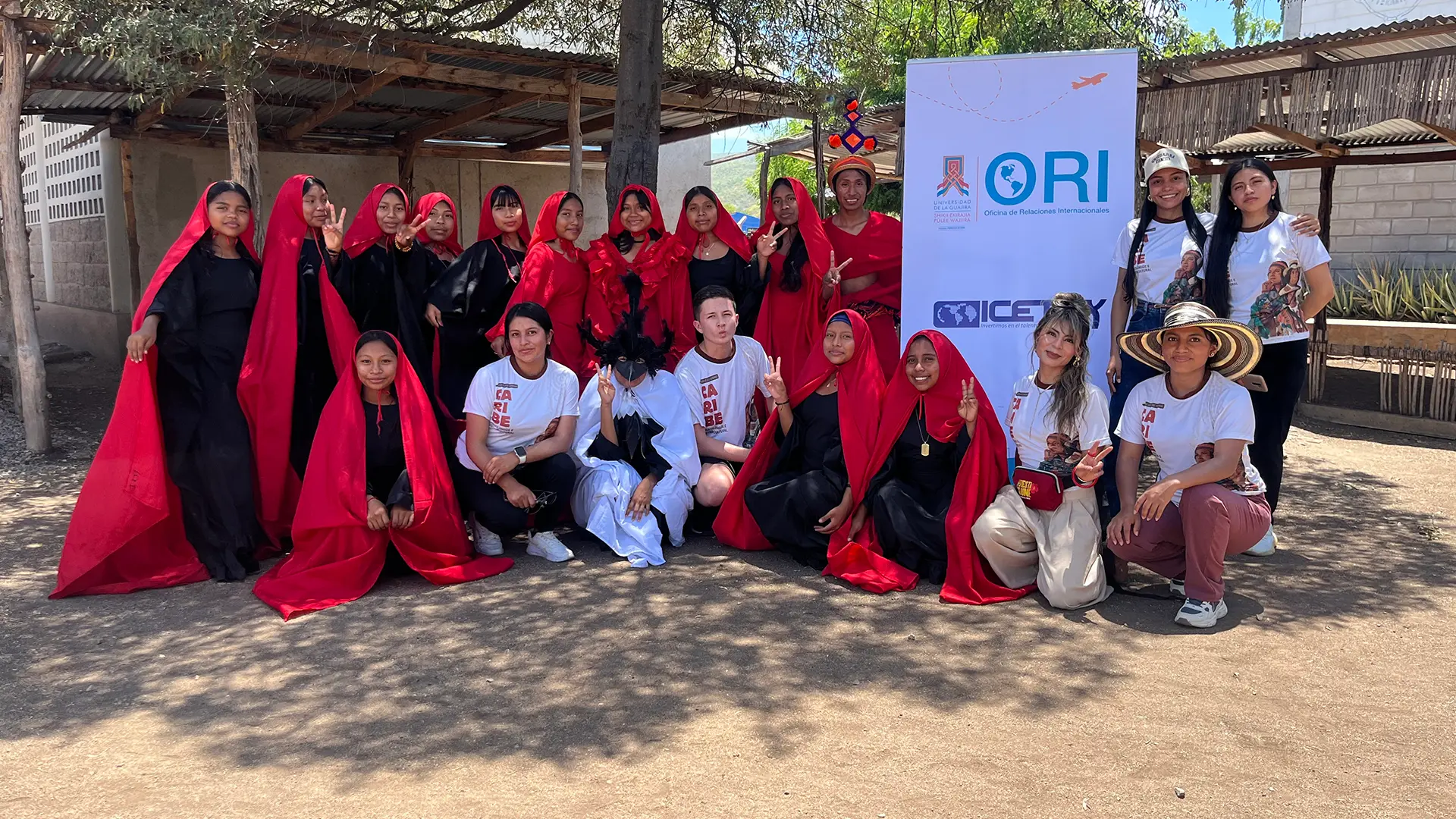 Grupo de personas reunidas al aire libre, algunas vestidas con atuendos tradicionales en colores rojo y negro, junto a representantes de la Oficina de Relaciones Internacionales (ORI). Al fondo se observa un aviso institucional y un entorno rural con árboles y construcciones tradicionales.