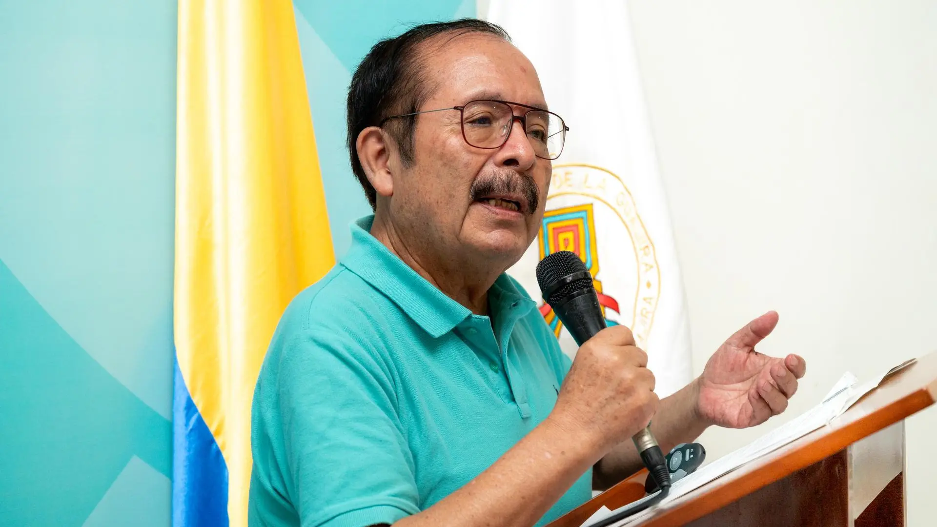 Un hombre adulto mayor, viste una camisa polo verde turquesa y habla sosteniendo un micrófono de mano mientras gesticula con la otra mano. Está junto a un atril de madera con papeles y un control remoto de presentaciones.
