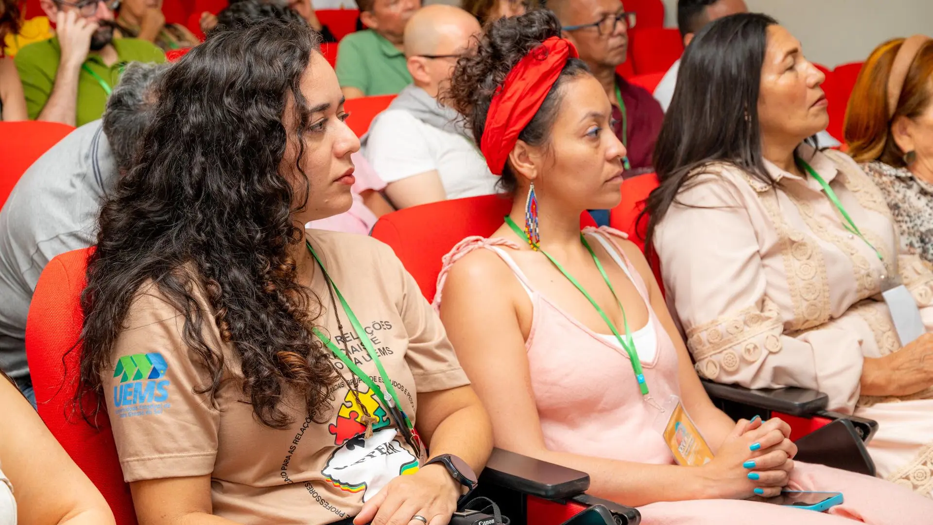 Tres mujeres sentadas en dentro de un auditorio miran con atención hacia el frente. La de la izquierda tiene el cabello largo y rizado, viste una camiseta beige y lleva una cámara Canon sobre las piernas. La del centro lleva un pañuelo rojo en la cabeza. La de la derecha tiene el cabello largo y oscuro y viste una blusa clara con encaje.