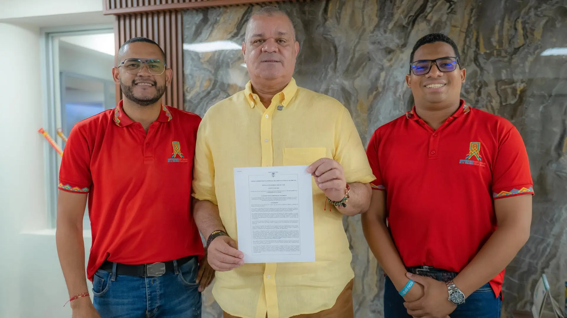 Tres hombres posan ante la cámara. El del centro, vestido con camisa amarilla, sostiene un documento mientras sonríe. A los lados, dos hombres con camisas rojas institucionales.