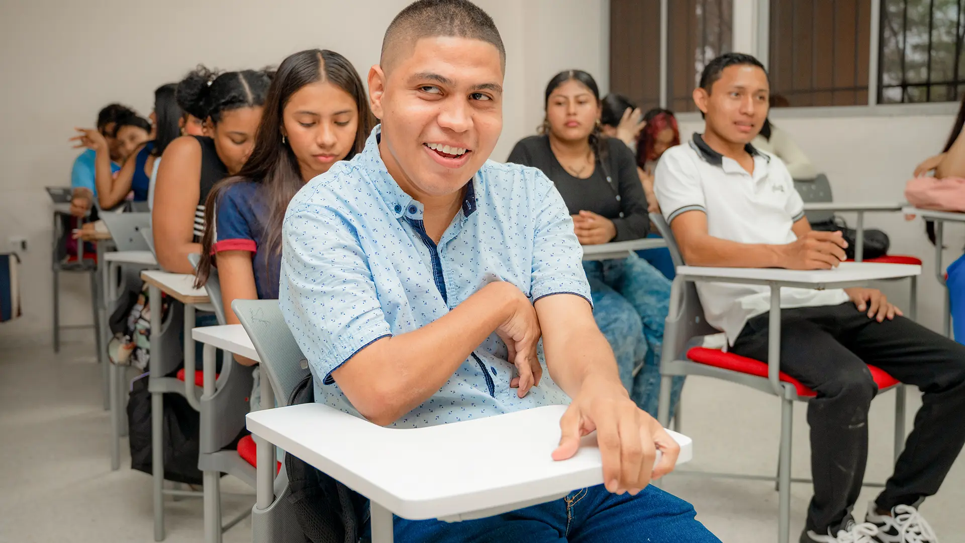 Rafael David Niño Atencio, estudiante de Trabajo Social de la Universidad de La Guajira, sonríe sentado en su pupitre durante una jornada académica, mientras sus compañeros permanecen en el aula.