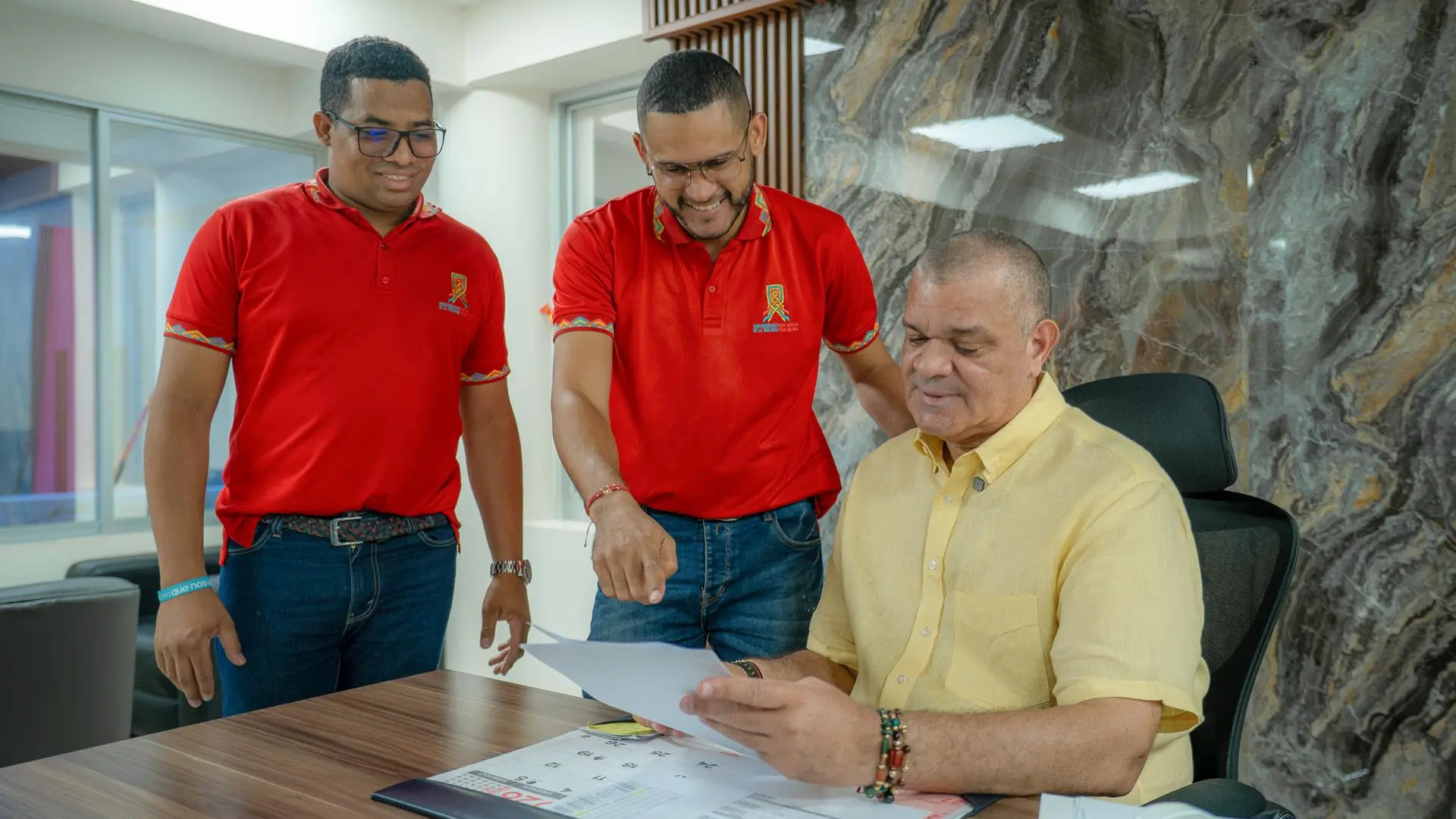 Tres hombres sonrientes, uno sentado y dos de pie visualizando un documento.