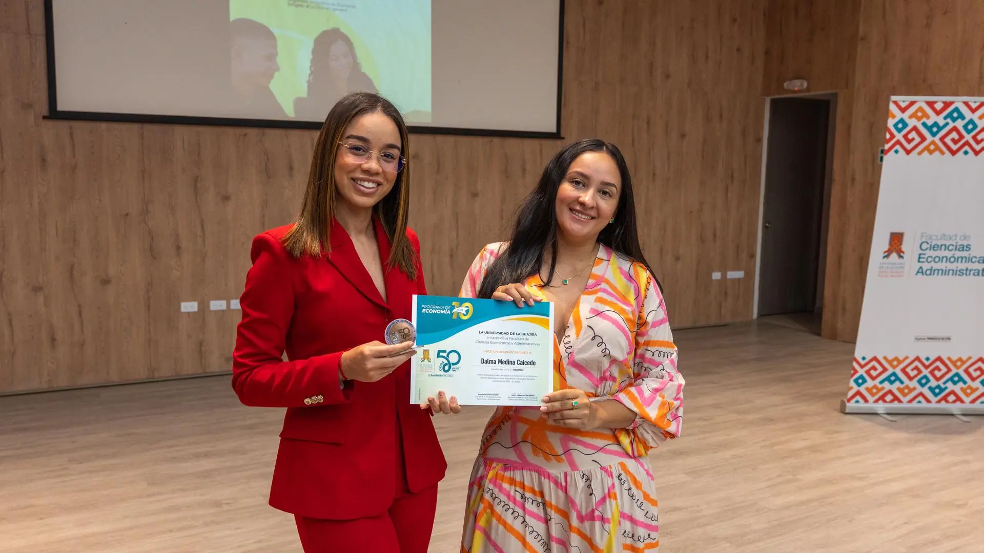 Dos mujeres sostienen un reconocimiento durante un evento académico de la Universidad de La Guajira. Al fondo se observan una pantalla de proyección y elementos institucionales en un auditorio.
